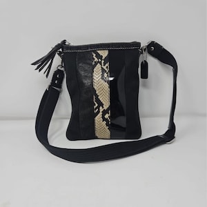 Może przedstawiać: Czarna torba na ramię z paskiem ze skóry węża pośrodku. Torba ma czarny regulowany pasek i małą czarną metkę. Torba ma czarny frędzel na zamku błyskawicznym.