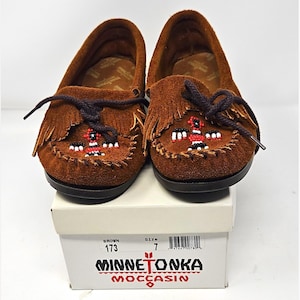 May include: A pair of brown suede moccasins with dark brown laces, fringe detailing, and a beaded bird design on the vamp. The shoes are resting on a white cardboard box labeled Minnetonka Moccasin with the text Brown 173 and Size 7 printed on it.