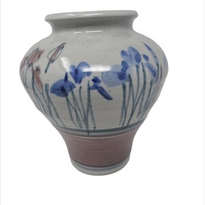May include: A ceramic vase with a wide, rounded body and a narrow base. The upper portion is off-white with hand-painted blue and muted pink floral motifs. The lower section features a solid, muted pink band above a white base rim.