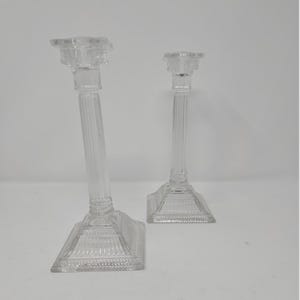 May include: Two clear glass candle holders. One is taller and leans slightly to the left. Both have a square base with a decorative pattern and a fluted column leading to a small, ornate candle cup.