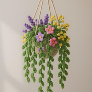Puede incluir: Una cesta colgante de ganchillo hecha a mano, llena de flores coloridas y hojas verdes colgantes. La cesta está suspendida por una cuerda beige, con detalles florales morados, rosas y amarillos. Una pieza decorativa encantadora.