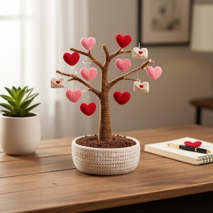 Puede incluir: Un árbol de ganchillo hecho a mano con tronco y ramas marrones, adornado con adornos de corazón rojos y rosas y pequeñas decoraciones de sobres blancos. El árbol está en una maceta de ganchillo blanca llena de tierra marrón, un objeto decorativo encantador.