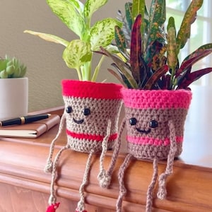 Może przedstawiać: Dwa szydełkowe doniczki z uśmiechniętymi twarzami i zwisającymi nogami. Jedna doniczka ma czerwoną obwódkę i pasek, druga różową obwódkę i pasek. Każda doniczka zawiera inną roślinę z zielonymi i czerwonymi liśćmi.