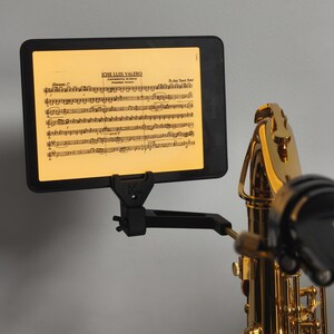 Op de afbeelding: Een zwarte tablet met bladmuziek, gemonteerd op een zwarte standaard, naast een gouden saxofoon. De tablet toont de naam "JOSE LUIS VALERO". De messing behuizing en het zwarte mondstuk van de saxofoon zijn zichtbaar.