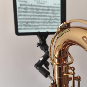 Puede incluir: Un saxofón dorado brillante con un brazo de montaje negro ajustable unido a su cuello. El brazo sostiene una tableta rectangular que muestra partituras musicales con la palabra FERVOR impresa en la parte superior de la pantalla en letra clara.