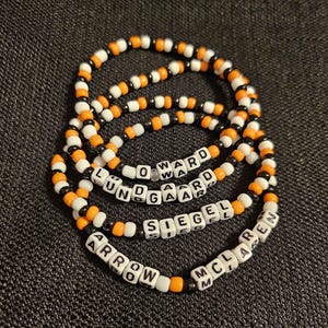 May include: Five black and orange beaded bracelets with white letter beads spelling out the names "Ward", "Lundgaard", "Siegel", "Arrow", and "McLaren".