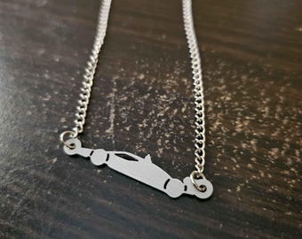 May include: A silver-colored necklace featuring a pendant shaped like a race car. The pendant is connected to a delicate chain. The necklace is displayed on a dark, textured surface.