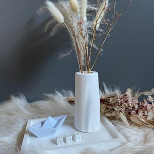 Könnte beinhalten: Weiße Keramikvase mit gerippter Textur, gefüllt mit getrocknetem Pampasgras und kleinen rosa Blüten. Die Vase steht auf einem weißen Tablett mit einem kleinen blauen Papierboot und weißen Buchstabenblöcken, die "TOM" buchstabieren.