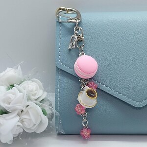 May include: A silver keychain charm featuring a pink macaron, a white teacup with brown liquid, and pink flower accents. The charm is attached to a light blue wallet.