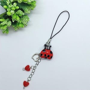 May include: A red and black ladybug charm with black dots hangs from a black string. The charm is attached to a silver chain with three red heart charms.