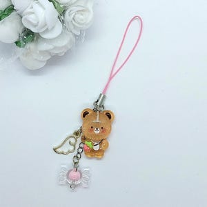 May include: A brown bear charm with a pink strap, a gold wing, and a pink candy charm. The bear has a white stripe on its head and is holding a strawberry. The charm is attached to a pink string.