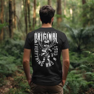 Puede incluir: Camiseta negra con un gr&aacute;fico blanco de un querub&iacute;n sosteniendo una ametralladora, alas y las palabras "BRANDED IN TYPE ORIGINAL EVERYTHING HAS A COST". La imagen muestra la espalda de una persona que lleva la camiseta en un entorno forestal.