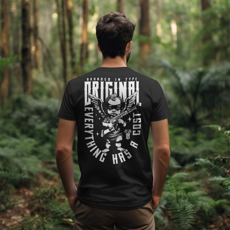 Puede incluir: Camiseta negra con un gr&aacute;fico blanco de un querub&iacute;n sosteniendo una ametralladora, alas y las palabras "BRANDED IN TYPE ORIGINAL EVERYTHING HAS A COST". La imagen muestra la espalda de una persona que lleva la camiseta en un entorno forestal.
