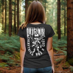 Puede incluir: Camiseta negra con un gr&aacute;fico blanco de un querub&iacute;n sosteniendo una ametralladora, con las palabras "ORIGINAL" y "EVERYTHING HAS A COST" en un dise&ntilde;o circular. La camiseta la lleva una persona en un entorno forestal.