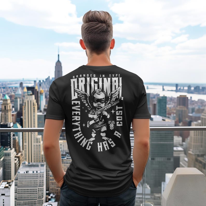 Puede incluir: Camiseta negra con un dise&ntilde;o gr&aacute;fico de un querub&iacute;n con un rifle, alas y las palabras "ORIGINAL" y "EVERYTHING HAS A COST". La persona lleva la camiseta con un paisaje urbano de fondo.