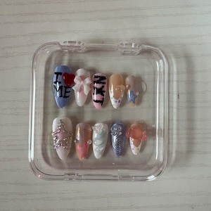 May include: A set of ten artificial nails in a clear plastic case. The nails feature various designs, including text, bows, and embellishments. Colors include blue, pink, orange, and white. Some nails have glitter and star accents.
