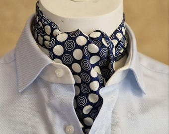 May include: A blue and white patterned ascot scarf is draped over a white mannequin head, set against a light blue button-down shirt. The scarf features a repeating pattern of white circles with concentric rings on a navy blue background.