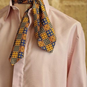 May include: A patterned ascot in shades of yellow, blue, and orange is draped over a pink dress shirt. The ascot features a geometric design. The shirt has a classic collar and buttoned cuffs. The background is a neutral color.