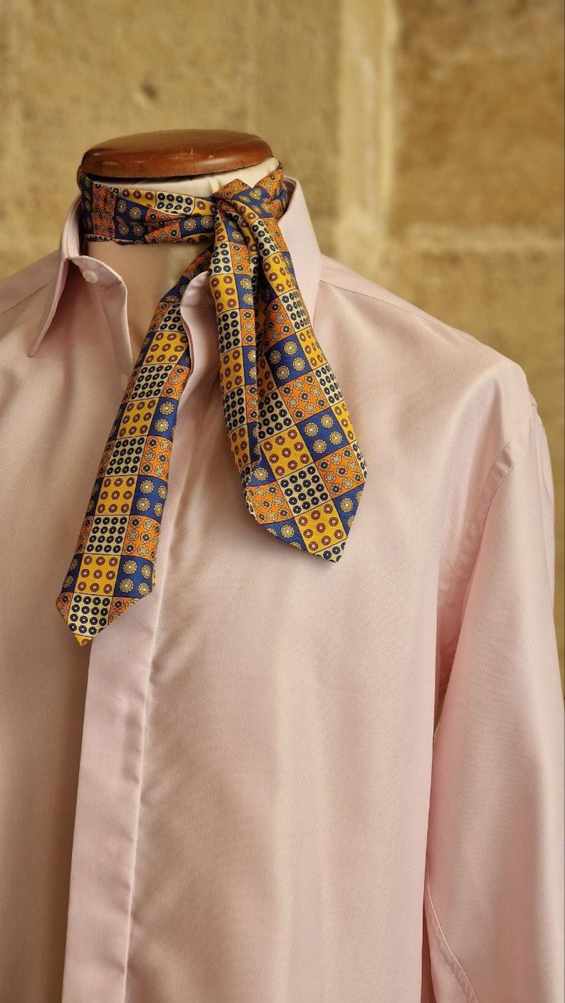 May include: A patterned ascot in shades of yellow, blue, and orange is draped over a pink dress shirt. The ascot features a geometric design. The shirt has a classic collar and buttoned cuffs. The background is a neutral color.