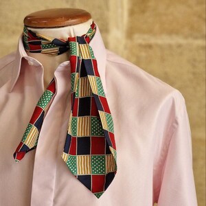 May include: A patterned necktie with a red, navy, green, and gold geometric design. The tie is knotted and worn over a light pink dress shirt. The tie's pattern includes squares, stripes, and small white stars.