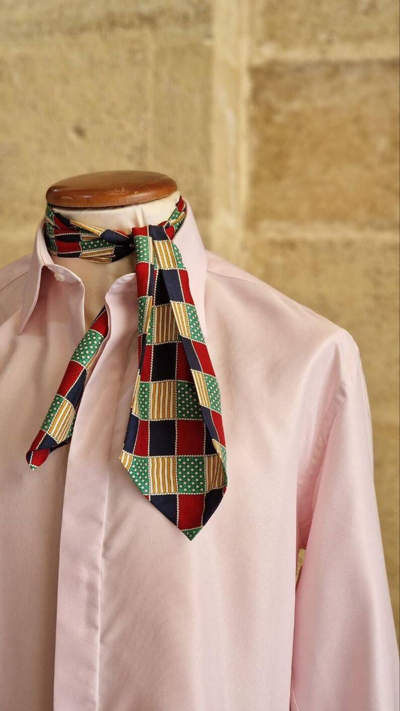 May include: A patterned necktie with a red, navy, green, and gold geometric design. The tie is knotted and worn over a light pink dress shirt. The tie's pattern includes squares, stripes, and small white stars.