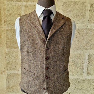 May include: A brown tweed vest with a notched lapel and a five-button closure. The vest has two front pockets and is displayed on a mannequin with a white dress shirt and a dark tie. The vest is a classic style.