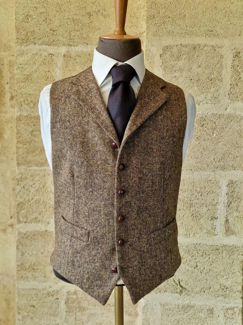 May include: A brown tweed vest with a notched lapel and a five-button closure. The vest has two front pockets and is displayed on a mannequin with a white dress shirt and a dark tie. The vest is a classic style.