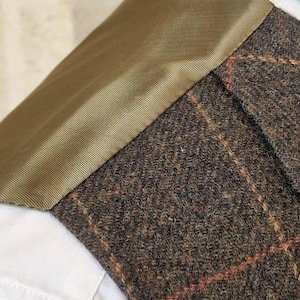 K&ouml;nnte beinhalten: Nahaufnahme eines Tweed-Jackenkragen mit braun-orangefarbenem Karomuster. Der Kragen ist mit olivgr&uuml;nem Stoff besetzt und sitzt auf einem wei&szlig;en Hemd.