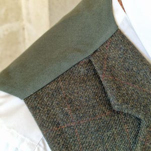 K&ouml;nnte beinhalten: Nahaufnahme einer Tweedweste mit gr&uuml;nem Kragen und einem wei&szlig;en Hemd mit Kragen darunter. Der Tweedstoff ist eine Mischung aus gr&uuml;nen, braunen und roten F&auml;den, die ein strukturiertes Muster erzeugen. Der Kragen der Weste ist einfarbig dunkelgr&uuml;n.