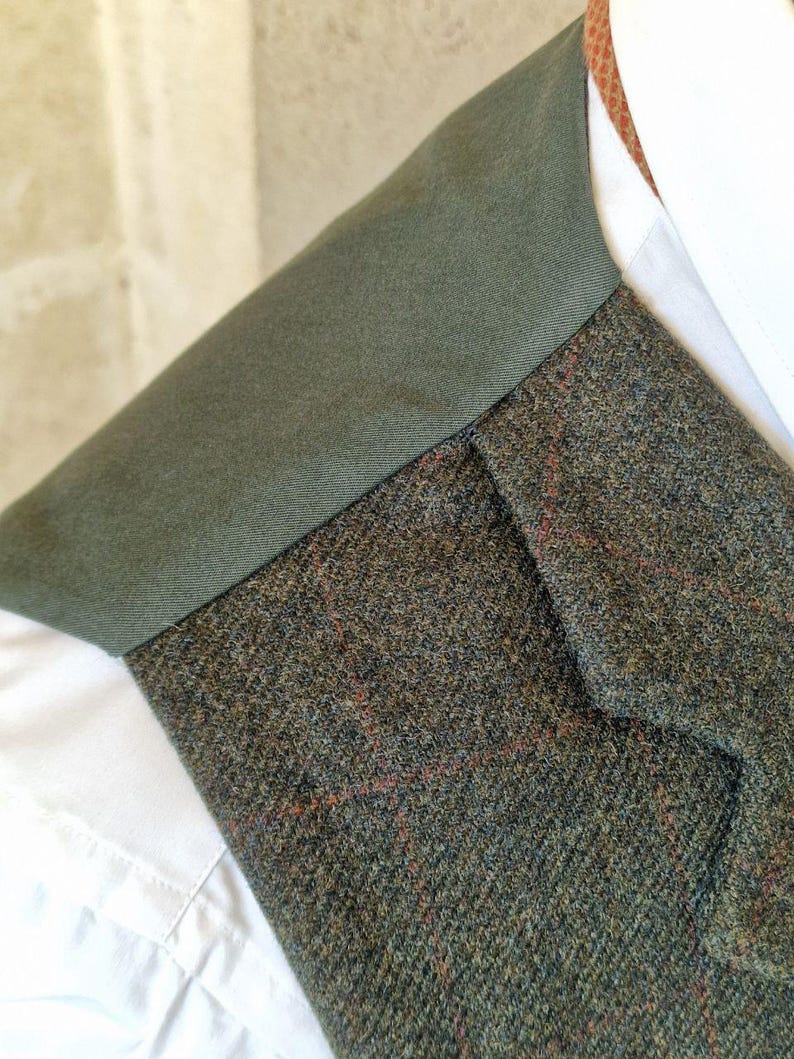 K&ouml;nnte beinhalten: Nahaufnahme einer Tweedweste mit gr&uuml;nem Kragen und einem wei&szlig;en Hemd mit Kragen darunter. Der Tweedstoff ist eine Mischung aus gr&uuml;nen, braunen und roten F&auml;den, die ein strukturiertes Muster erzeugen. Der Kragen der Weste ist einfarbig dunkelgr&uuml;n.