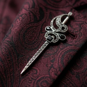 Può includere: Spilla da bavero argentata con un design ornato con dettagli neri e una pietra nera quadrata. La spilla è posta su un tessuto bordeaux a fantasia paisley, creando un contrasto sorprendente. Il design include una parte superiore decorativa e un lungo stelo appuntito.