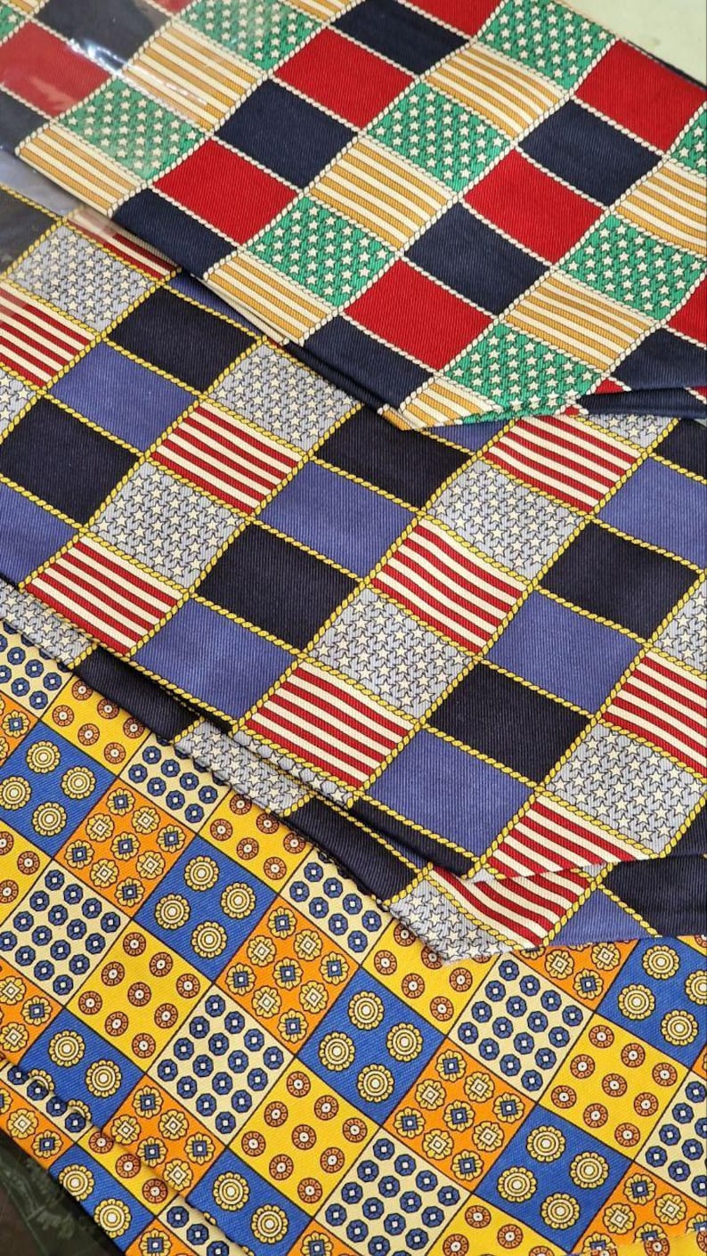 May include: Close-up of several patterned neckties. The ties feature a variety of designs, including red and white stripes, blue and white stars, and geometric patterns in yellow, blue, and red. The ties are arranged in a layered fashion.