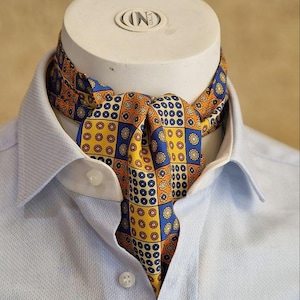 Puede incluir: Un pañuelo de seda estampado en tonos amarillos, naranjas y azules, usado con una camisa de cuello azul claro. El pañuelo presenta un diseño geométrico repetitivo. El pañuelo se muestra en una cabeza de maniquí blanca.