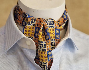 May include: A patterned silk ascot in shades of yellow, orange, and blue, worn with a light blue collared shirt. The ascot features a repeating geometric design. The ascot is displayed on a white mannequin head.