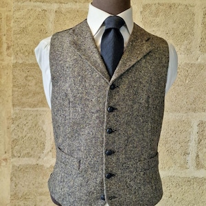 May include: A tweed vest in shades of brown, gray, and green, worn over a white dress shirt and a dark blue tie. The vest has a classic, tailored fit with black buttons and two front pockets. The outfit is displayed on a mannequin.