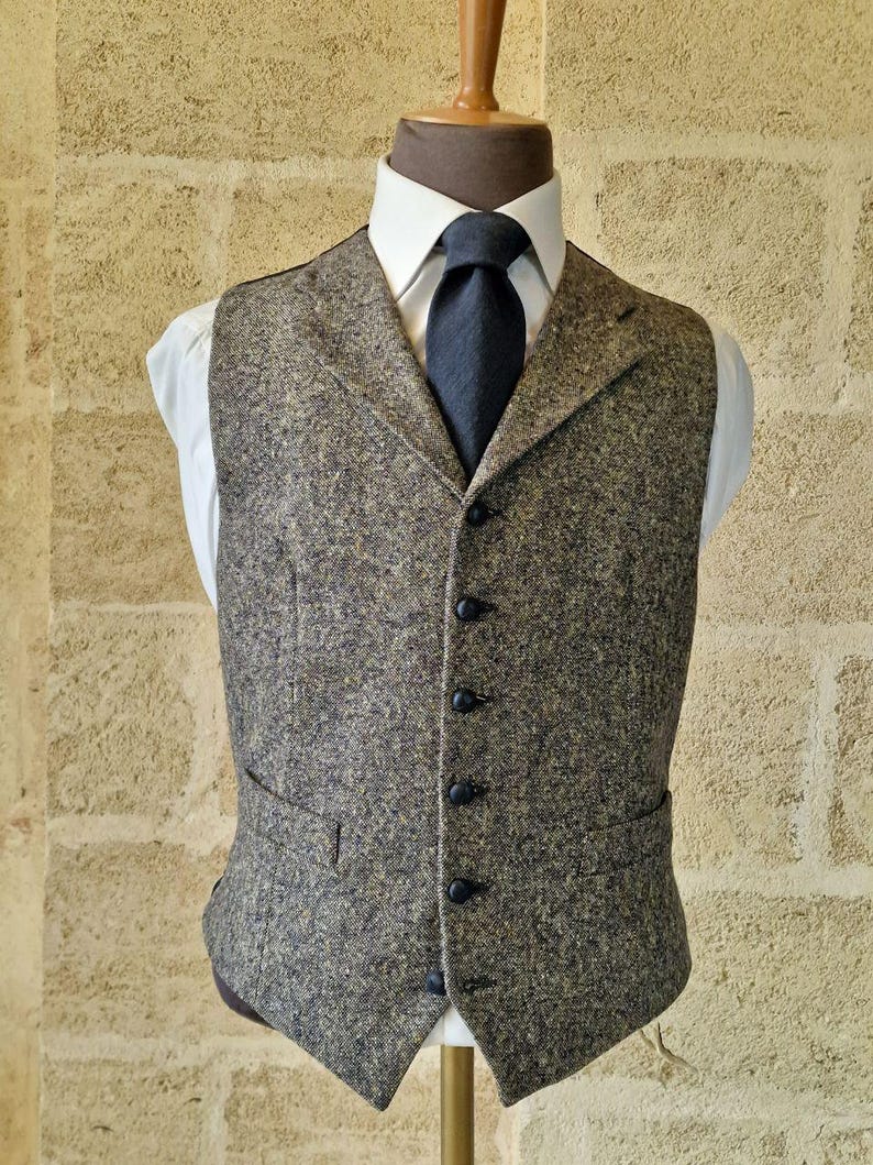 May include: A tweed vest in shades of brown, gray, and green, worn over a white dress shirt and a dark blue tie. The vest has a classic, tailored fit with black buttons and two front pockets. The outfit is displayed on a mannequin.