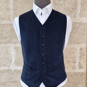 Pode incluir: Um colete azul marinho escuro com fecho de seis botões e dois bolsos frontais. É usado sobre uma camisa branca com gola clássica. O colete é exibido em um manequim contra um fundo texturizado marrom claro.