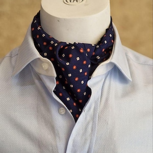 May include: A navy ascot with a small floral pattern of red and white flowers is worn with a light blue button-down shirt. The ascot is displayed on a white mannequin head.