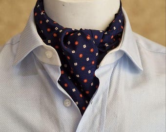 May include: A navy ascot with a small floral pattern of red and white flowers is worn with a light blue button-down shirt. The ascot is displayed on a white mannequin head.