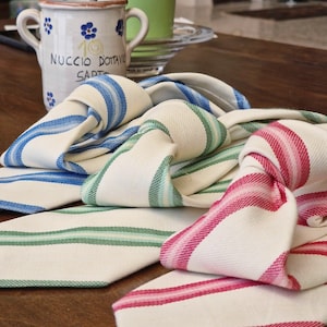 May include: Three neckties made of off-white fabric with woven striped patterns in blue, green, and pink. Each tie features a pointed end and a knotted section, displaying a textured weave. The ties are arranged overlapping on a wooden surface.