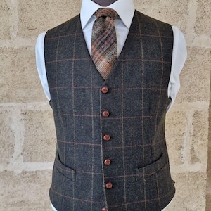 Può includere: Un gilet in tweed grigio scuro con motivo a quadri, bottoni marroni e due tasche anteriori. Il gilet è indossato sopra una camicia bianca e abbinato a una cravatta fantasia. L'indumento è esposto su un manichino.