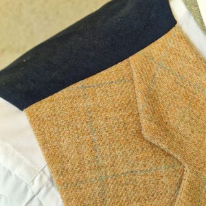 K&ouml;nnte beinhalten: Nahaufnahme einer beigefarbenen Tweedweste mit marineblauem Kragen. Die Weste hat ein dezentes blaues Karomuster und wird &uuml;ber einem wei&szlig;en Hemd mit Kragen getragen. Der Stoff hat eine strukturierte Optik.