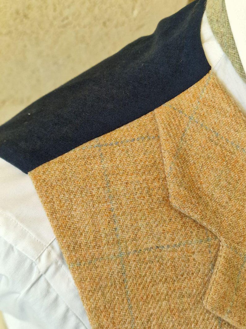 K&ouml;nnte beinhalten: Nahaufnahme einer beigefarbenen Tweedweste mit marineblauem Kragen. Die Weste hat ein dezentes blaues Karomuster und wird &uuml;ber einem wei&szlig;en Hemd mit Kragen getragen. Der Stoff hat eine strukturierte Optik.