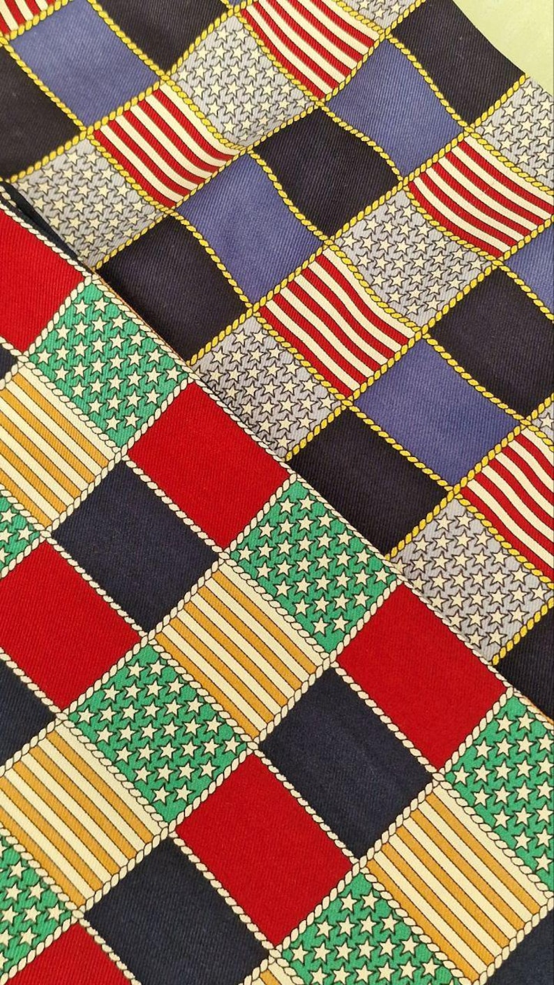 May include: Close-up of patterned fabric featuring a repeating design of squares. The squares display patriotic motifs: red and white stripes, white stars on blue, and alternating solid colors. The squares are outlined with a gold rope-like trim.
