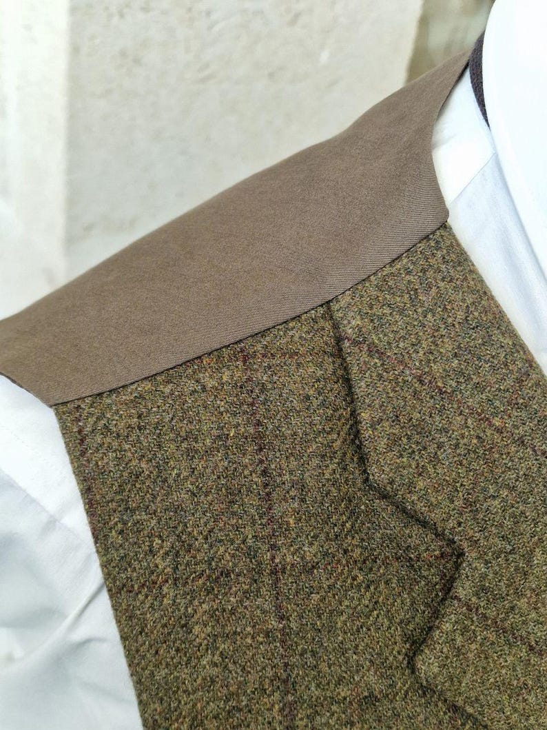 K&ouml;nnte beinhalten: Nahaufnahme einer Tweedweste mit braunem Kragen. Die Weste besteht aus einer Mischung aus gr&uuml;nen, braunen und roten F&auml;den, die ein strukturiertes Aussehen erzeugen. Ein wei&szlig;es Hemd ist darunter zu sehen, was auf formelle oder Business-Kleidung hindeutet.
