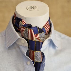 May include: A blue ascot with a patchwork design featuring red and white stripes and stars, worn with a light blue button-down shirt. The ascot is knotted at the neck, showcasing a patriotic theme.