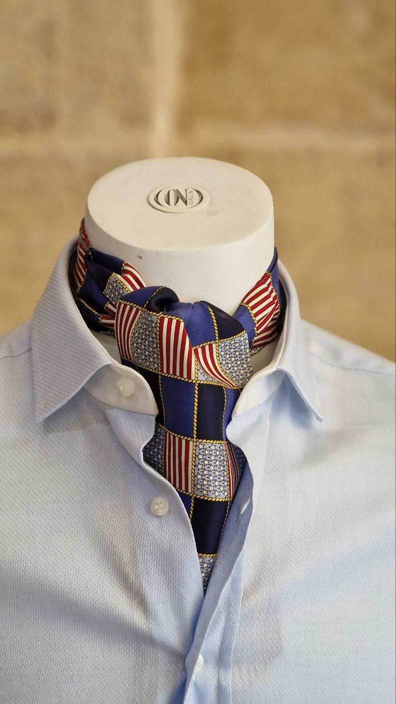 May include: A blue ascot with a patchwork design featuring red and white stripes and stars, worn with a light blue button-down shirt. The ascot is knotted at the neck, showcasing a patriotic theme.