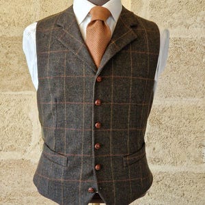 K&ouml;nnte beinhalten: Eine braune Tweedweste mit Karomuster, kombiniert mit einem wei&szlig;en Hemd und einer rostfarbenen Krawatte. Die Weste hat ein klassisches Design mit fallendem Revers, zwei Vordertaschen und Holzkn&ouml;pfen. Das Outfit wird auf einer Schaufensterpuppe pr&auml;sentiert.