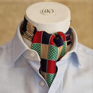 May include: A colorful ascot scarf with a patchwork design of red, green, navy, and tan squares, worn with a light blue button-down shirt. The scarf is displayed on a white mannequin head.