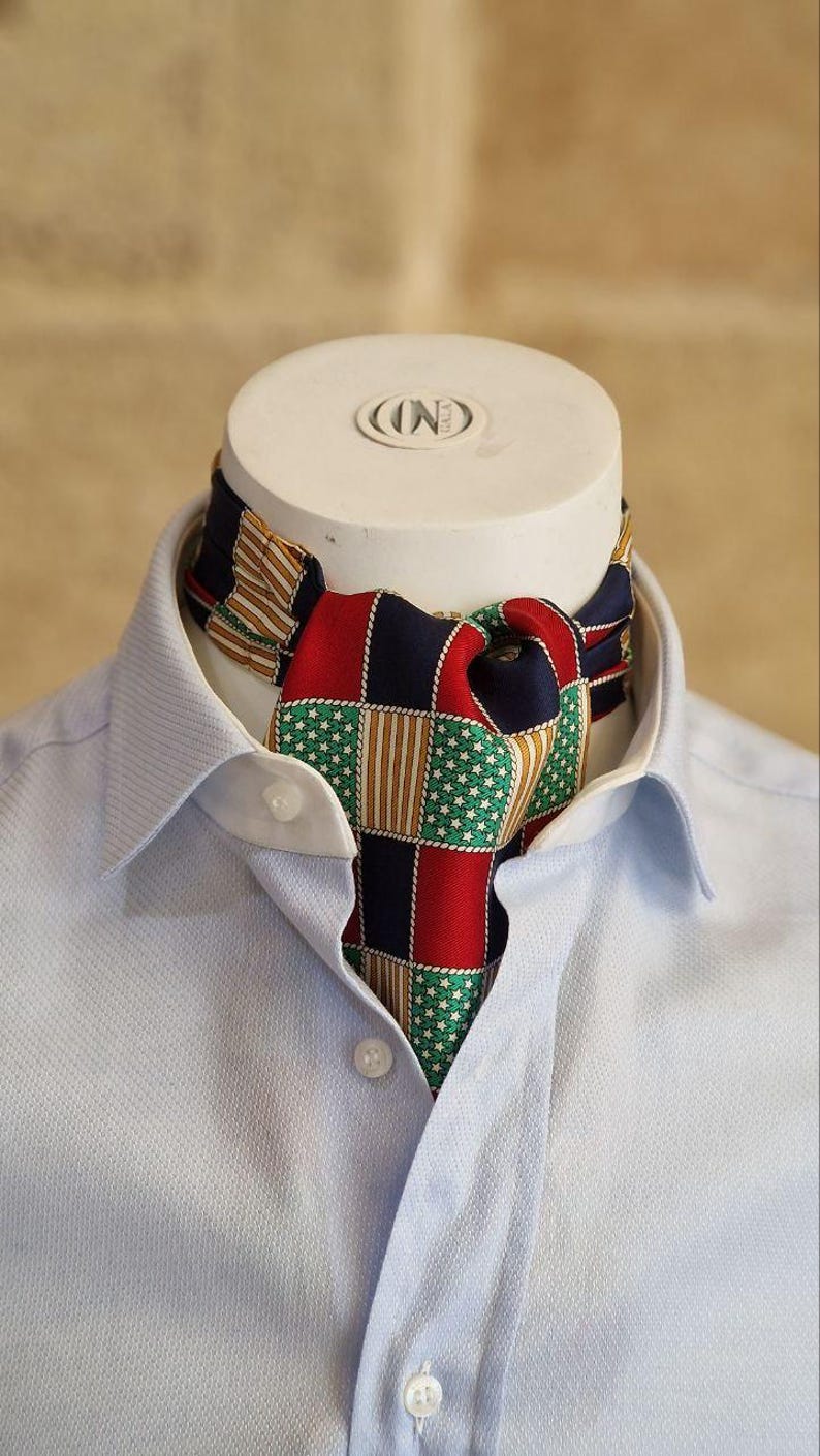 May include: A colorful ascot scarf with a patchwork design of red, green, navy, and tan squares, worn with a light blue button-down shirt. The scarf is displayed on a white mannequin head.